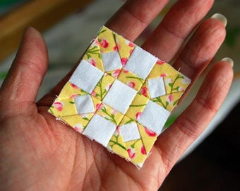 May include: A small, square patchwork quilt block held in a hand. The block features a yellow floral print fabric with white squares arranged in a grid pattern. The floral print includes pink flowers and green leaves.