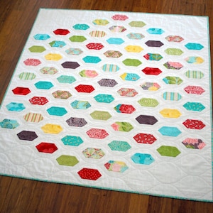 May include: A white quilt with a diamond-shaped pattern of colorful fabric hexagons. The hexagons are arranged in rows, with each row offset from the one above it. The quilt is made with a variety of fabrics, including floral prints, stripes, and solids.