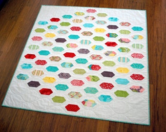 May include: A white quilt with a diamond-shaped pattern of colorful fabric hexagons. The hexagons are arranged in rows, with each row offset from the one above it. The quilt is made with a variety of fabrics, including floral prints, stripes, and solids.