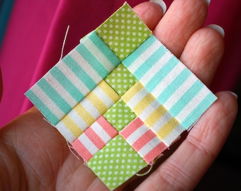 May include: A small, square fabric patchwork with a woven design. The patchwork features alternating stripes and polka dots in pastel colors: light blue, yellow, pink, and green. The fabric is held in a hand.