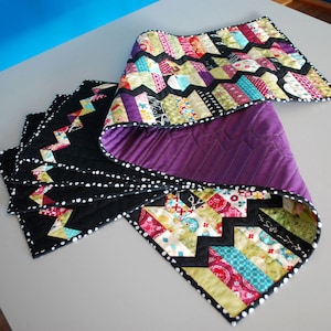 May include: A colourful table runner with a chevron pattern in shades of pink, green, blue, and yellow. The runner is edged with black fabric with white polka dots. The back of the runner is purple.