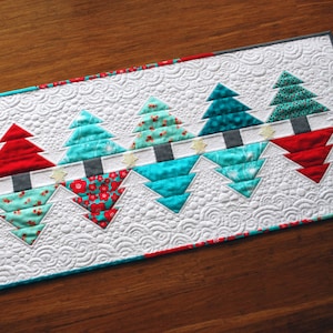 May include: A white quilted table runner with a row of turquoise and red Christmas trees. The trees are outlined in white and have a white star at the base.