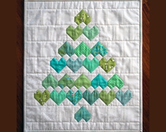 May include: A white quilted fabric square with a green and blue patchwork heart-shaped Christmas tree design.