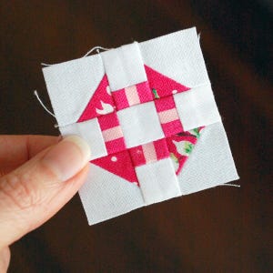 May include: A small, square quilt block featuring a central design of pink and white fabric. The pink fabric has a floral pattern and is arranged in a geometric pattern. The block is held by a hand.