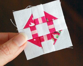 May include: A small, square quilt block featuring a central design of pink and white fabric. The pink fabric has a floral pattern and is arranged in a geometric pattern. The block is held by a hand.