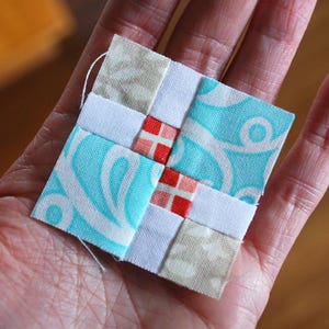 May include: A small, square fabric patchwork piece with a cross design. The patchwork features light blue, white, beige, and red squares. The light blue squares have a white swirl pattern. A white thread is attached to the top.
