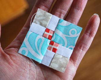May include: A small, square fabric patchwork piece with a cross design. The patchwork features light blue, white, beige, and red squares. The light blue squares have a white swirl pattern. A white thread is attached to the top.