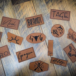 May include: A collection of 14 brown leather patches with black lettering and designs. The patches feature names, initials, and images like a bull skull, a fish, and a mountain range.