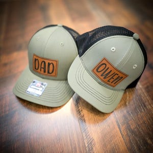 May include: Two olive green and black trucker hats with brown leather patches. One hat has the word "DAD" and the other has "OWEN" in brown lettering. The hats are on a wooden surface.