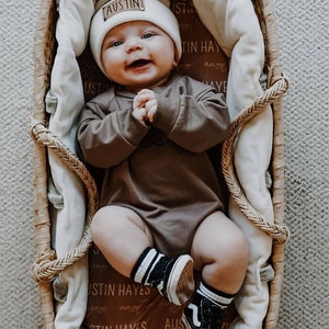 May include: A baby wearing a brown onesie, white beanie with the name "Austin" on it, and black and white striped socks. The baby is lying in a wicker basket with a white blanket.