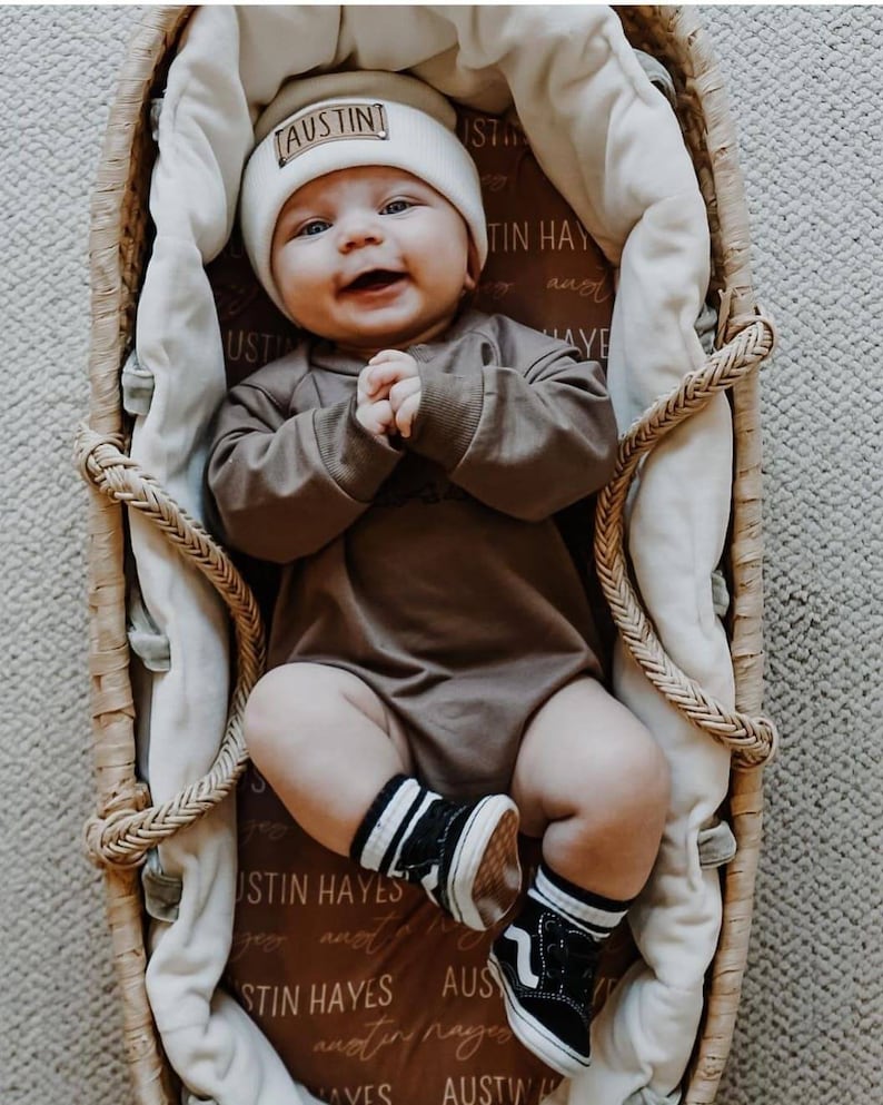 May include: A baby wearing a brown onesie, white beanie with the name "Austin" on it, and black and white striped socks. The baby is lying in a wicker basket with a white blanket.