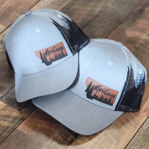 May include: Two gray and black trucker hats with a brown leather patch that says "Montana" and a silhouette of a pine forest.