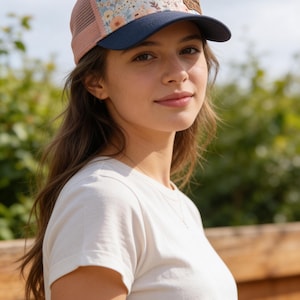 May include: A floral print trucker hat with a navy blue brim and a tan leather patch. The hat features a mesh back and a pink side panel. The hat is worn by a person wearing a white t-shirt.