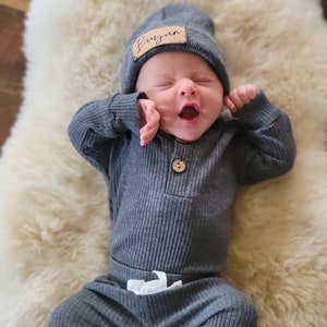 May include: A baby wearing a gray ribbed knit onesie and matching pants with a white drawstring. The baby is wearing a gray beanie with a brown leather name tag that says "Bryan".