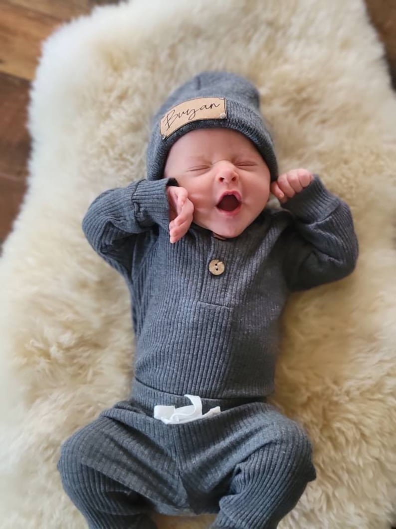 May include: A baby wearing a gray ribbed knit onesie and matching pants with a white drawstring. The baby is wearing a gray beanie with a brown leather name tag that says "Bryan".