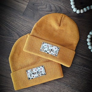 May include: Two mustard yellow knit beanies with personalized floral patches. The top beanie has the name "Junie" on the patch and the bottom beanie has the name "Poppy" on the patch.