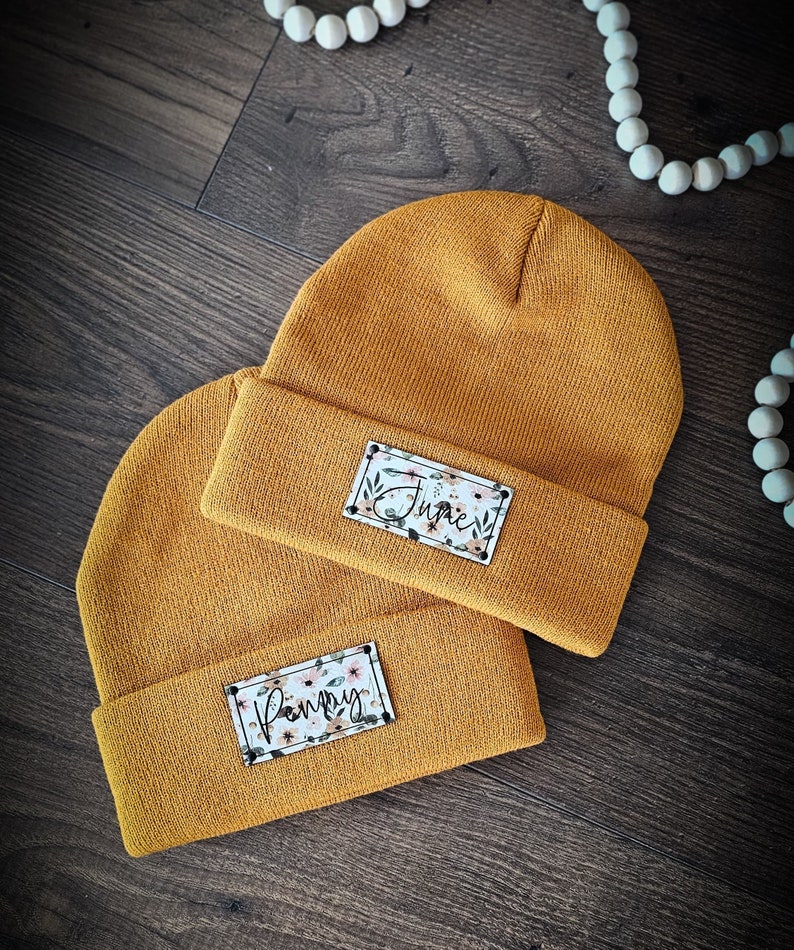 May include: Two mustard yellow knit beanies with personalized floral patches. The top beanie has the name "Junie" on the patch and the bottom beanie has the name "Poppy" on the patch.