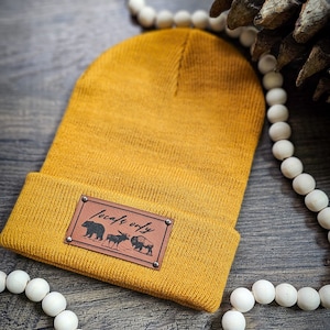 May include: Mustard yellow beanie with a brown leather patch. The patch features the text "Locals Only" above illustrations of a bear, moose, and bison. The hat is folded at the brim and rests on a wooden surface.