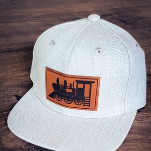 May include: A gray and white baseball cap with a brown leather patch featuring a black silhouette of a train engine.