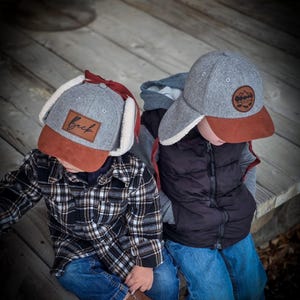 May include: Two children wearing gray and brown baseball caps with ear flaps. One cap has the word "Beck" on it. The other cap has a mountain and tree logo. One child is wearing a black and white plaid shirt and the other is wearing a black vest.