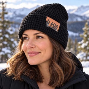 May include: A black waffle-knit beanie featuring a tan leather patch on the front. The patch is shaped like the state of Montana and displays a black forest silhouette graphic with the number 406 printed in the center, secured by two rivets.