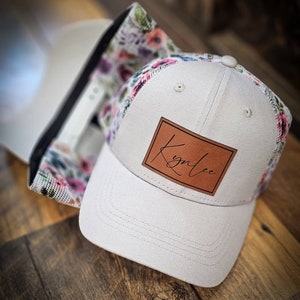 May include: A white and grey baseball cap with a floral mesh back. The cap has a brown leather patch with the name "Kynlee" written in cursive.