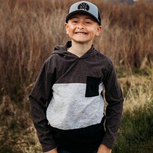 Peut inclure: Un jeune garçon portant un sweat à capuche noir et gris avec une poche noire, un jean bleu et une casquette de baseball bleue et noire avec un logo blanc qui dit "MXB".