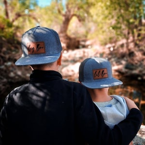 Peut inclure: Deux enfants portant des casquettes de baseball bleues et grises avec des patchs en cuir marron. Le patch avant sur la plus grande casquette dit "Big Bro" et la plus petite casquette dit "Lil Bro".