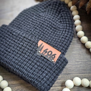 May include: A grey knitted beanie with a brown leather patch featuring a silhouette of trees and the number "406".