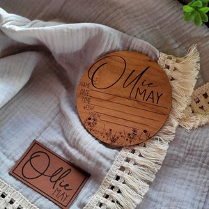 May include: A light blue muslin baby blanket with a white crocheted trim. The blanket has two wooden plaques with the text "Ollie May" on them. One plaque is round and has a floral design. The other plaque is rectangular and has a simple design.