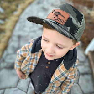 May include: A young child wearing a camouflage trucker hat with a brown leather patch that says "T-Rex" and a black dinosaur skeleton graphic. The child is also wearing a plaid shirt over a black shirt with white stars and brown boots.