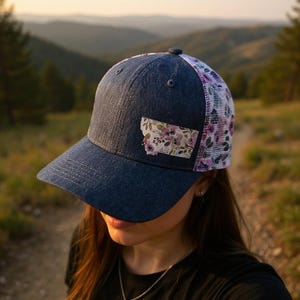 May include: A navy blue baseball cap with a floral Montana state patch. The cap has a mesh back with a purple floral pattern and a curved peak. The hat is worn outdoors with a scenic mountain backdrop.