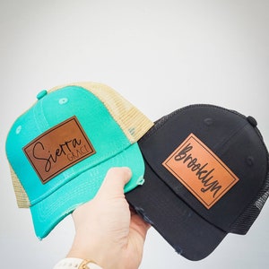 May include: Two trucker hats, one teal and one black, each with a distressed brim and a leather patch. The teal hat has "Sierra Grace" on the patch, and the black hat has "Brooklyn". Both hats have a mesh back.