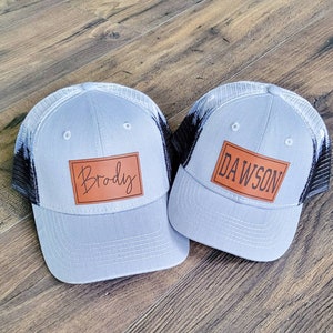May include: Two gray and black trucker hats with brown leather patches. The patch on the left hat says "Brody" and the patch on the right hat says "Dawson".