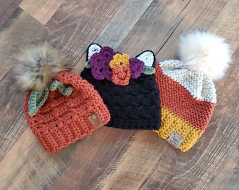Puede incluir: Tres gorros de punto con diferentes diseños. Un gorro es naranja y tiene un diseño de hoja verde, otro es negro con un diseño de flor y el último es a rayas de naranja, blanco y amarillo.