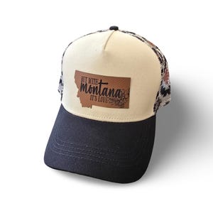 May include: A trucker hat with a black brim and a cream-colored front panel. A brown leather-like patch in the shape of Montana is centered on the front, featuring the black text "BUT WITH montana IT'S LOVE" and a floral graphic design.