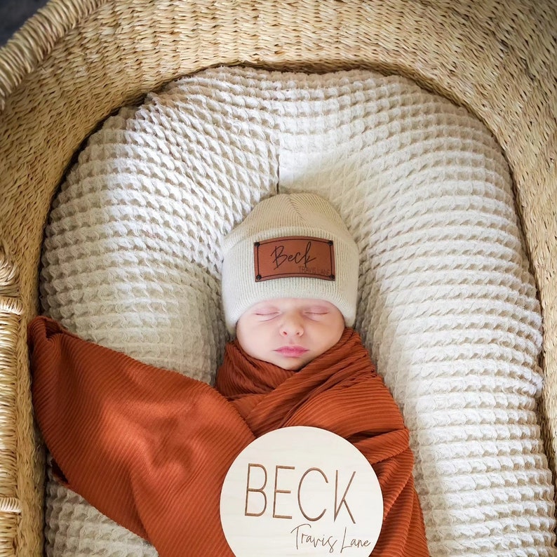 May include: A newborn baby boy is sleeping in a woven basket with a brown leather name tag on a cream beanie that says "Beck" and a wooden sign that says "BECK Travis Lane".