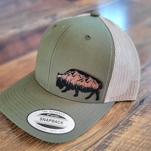 May include: A green and beige trucker hat with a black silhouette of a buffalo standing in front of a mountain range with pink peaks. The hat has a brown leather patch with the text "THE AUTHENTIC YUPOONG SNAPBACK YUPOONG SINCE 1974".