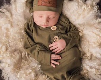 May include: A newborn baby wearing a green knotted hat with a leather patch that says "Bodhi" and a green knotted gown with wooden buttons.