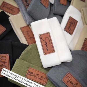 May include: A variety of beanies with personalized leather patches. The beanies are in different colors, including gray, white, green, and black. The patches have names written on them, such as "Sienna", "Bentley", "Nealy", and "Jack".
