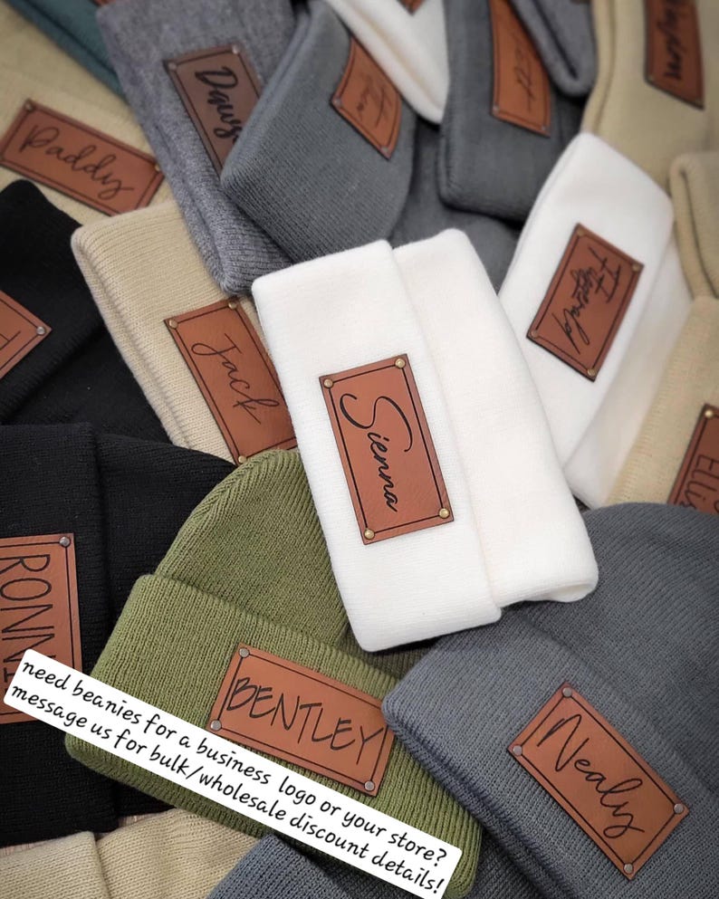 May include: A variety of beanies with personalized leather patches. The beanies are in different colors, including gray, white, green, and black. The patches have names written on them, such as "Sienna", "Bentley", "Nealy", and "Jack".