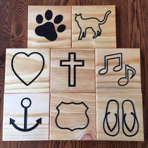 May include: Eight square wooden tiles featuring black engraved icons including a paw print, a cat silhouette, a heart, a cross, musical notes, an anchor, a shield-shaped emblem, and a pair of flip-flops, all displaying natural wood grain.