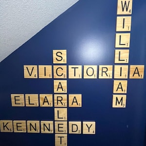 May include: A wall display of light-colored wooden square tiles featuring black letters and numerical values, arranged in a crossword-style pattern to spell out the names Victoria, William, Scarlett, Elara, and Kennedy in a vertical and horizontal layout.