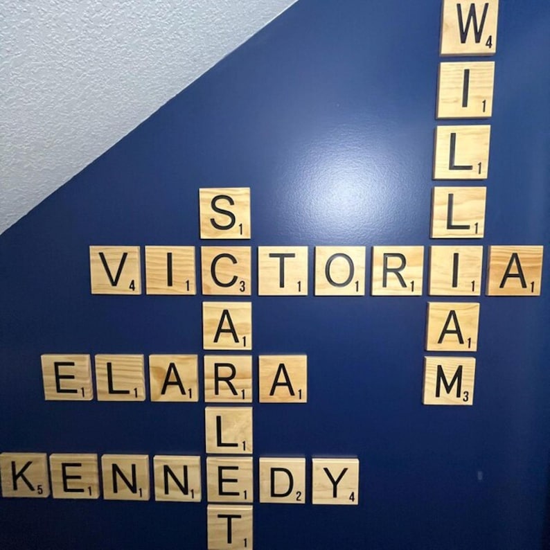 May include: A wall display of light-colored wooden square tiles featuring black letters and numerical values, arranged in a crossword-style pattern to spell out the names Victoria, William, Scarlett, Elara, and Kennedy in a vertical and horizontal layout.