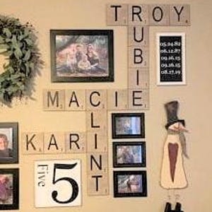 May include: A wall display featuring wooden letter tiles arranged in a crossword pattern spelling TROY, RUBIE, MACIE, KARLINT, and the number 5. The arrangement includes several framed photographs, a vertical sign with five dates, and a snowman cutout.