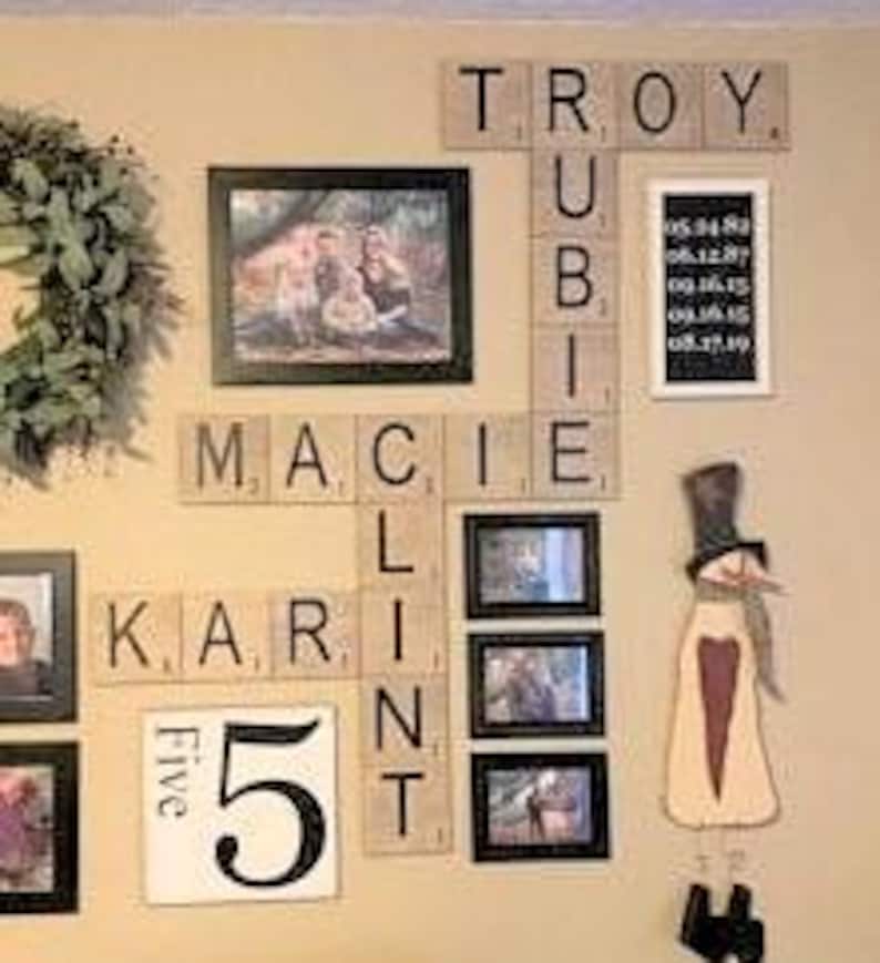 May include: A wall display featuring wooden letter tiles arranged in a crossword pattern spelling TROY, RUBIE, MACIE, KARLINT, and the number 5. The arrangement includes several framed photographs, a vertical sign with five dates, and a snowman cutout.