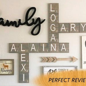 May include: A wall display featuring wooden tiles arranged like a crossword puzzle with the names LOGAN, GARY, ALAINA, and LEXI. The tiles are grey with white letters and small numerical values. A black cursive word family is mounted above the tiles.