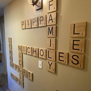 May include: A wall display of large, square wooden tiles resembling Scrabble pieces, arranged in a crossword pattern. The tiles feature black letters and numerical values, spelling out names and words including PIPPA, FAMILY, NICHOLAS, LEE, HOPE, and EFFORT.
