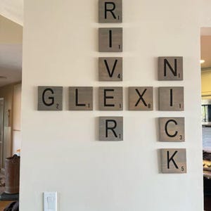 May include: A wall-mounted display of square wooden tiles featuring black letters and numerical values, arranged in a crossword-style pattern to spell the names River, Lexi, and Nick, with a grey wood grain finish on each individual tile piece.