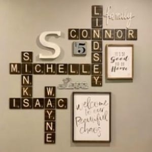 May include: A wall display featuring dark brown wooden tiles with white letters arranged like a crossword puzzle, spelling names including Michelle, Isaac, Wayne, Lindsey, and Connor. It includes a large white letter S, a calendar tile, and two framed signs.